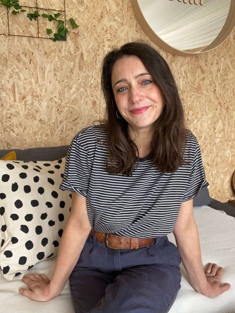 Woman sitting on a sofa with a striped tshirt on. She is smiling at the camera. Theres a spotted cushion behind her and the background is a wood chip wall