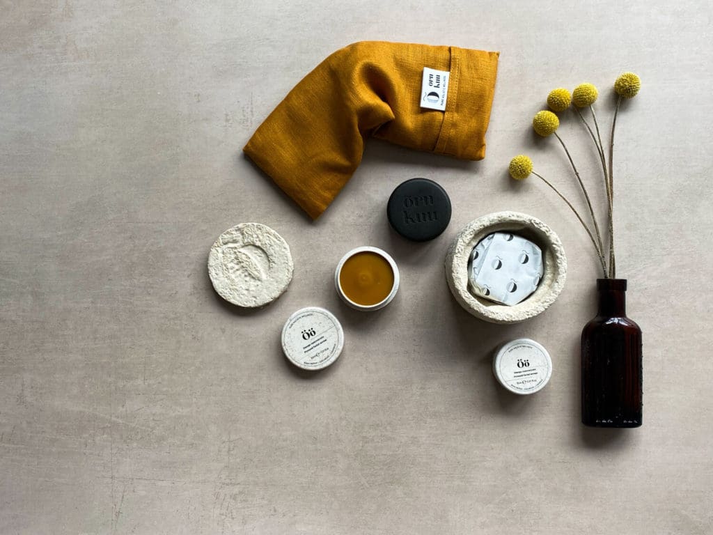 The picture shows the products available on the website. There are product jars and a bottle arranged on a grey beige backdrop and a mustard yellow linen eye pillow